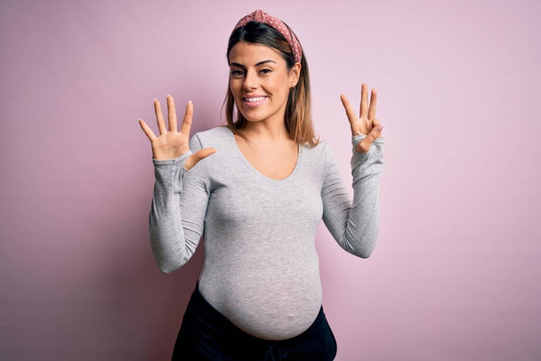 Young beautiful brunette woman pregnant expecting baby over isolated pink background showing and pointing up with fingers number eight while smiling confident and happy. - Foto, Imagem