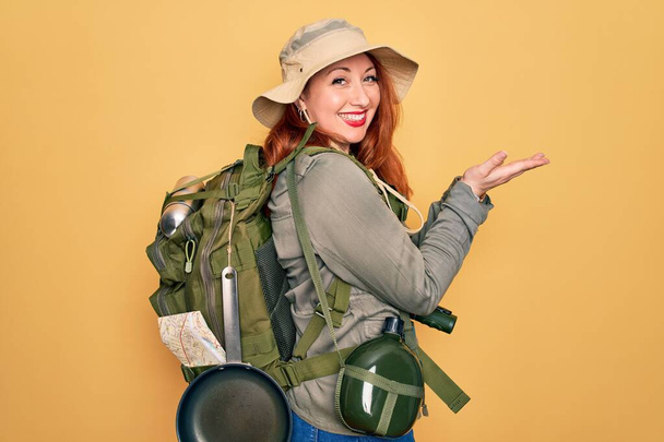 Young redhead backpacker woman hiking wearing backpack and hat over yellow background pointing aside with hands open palms showing copy space, presenting advertisement smiling excited happy - Photo, Image