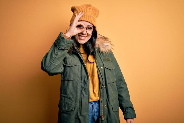 Молода брюнетка в окулярах і зимове пальто з капелюхом над жовтим ізольованим тлом робить нормальний жест з посмішкою руки, очі дивиться крізь пальці з щасливим обличчям
. - Фото, зображення