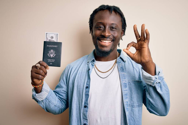 Νεαρός Αφροαμερικάνος τουρίστας που κατέχει διαβατήριο με χαρτονομίσματα δολαρίων, όπως τα χρήματα ταξιδεύουν κάνει ok υπογράψει με τα δάχτυλα, εξαιρετικό σύμβολο - Φωτογραφία, εικόνα