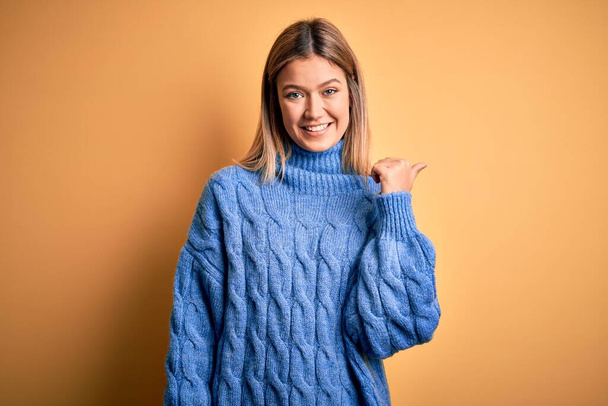 Young beautiful blonde woman wearing turtleneck sweater over yellow isolated background smiling with happy face looking and pointing to the side with thumb up. - Photo, image