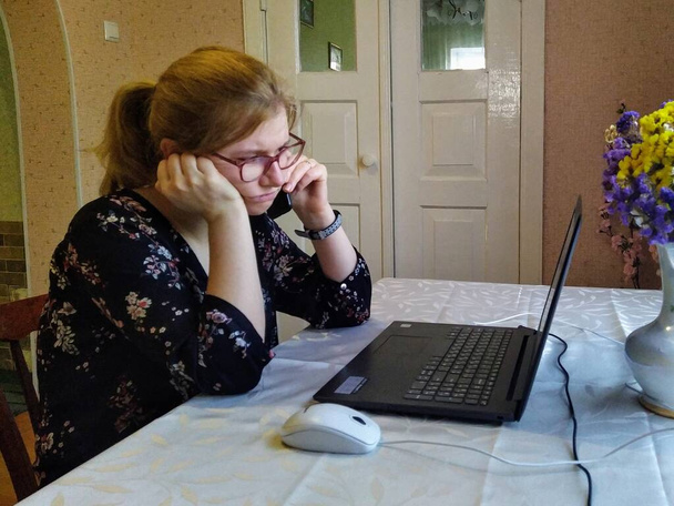 不満なヨーロッパの若い女性はテーブルに座って、スマートフォンで話し、自宅でノートパソコンで作業します - 写真・画像