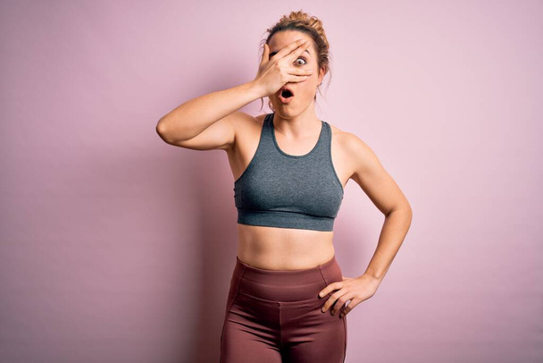 Νεαρή όμορφη ξανθιά αθλήτρια που κάνει αθλητισμό φορώντας αθλητικά ρούχα πάνω από ροζ φόντο κρυφοκοιτάζοντας σε σοκ που καλύπτει το πρόσωπο και τα μάτια με το χέρι, κοιτάζοντας μέσα από τα δάχτυλα με αμηχανία έκφραση. - Φωτογραφία, εικόνα
