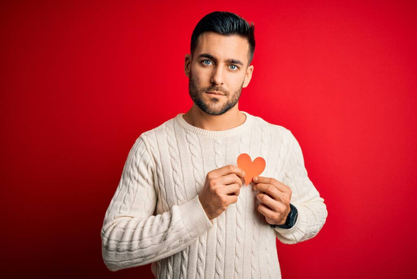 Młody przystojny mężczyzna trzyma czerwony kształt serca jako symbol romantyczny i zdrowia z pewnością wyraz na inteligentnej twarzy myśląc poważnie - Zdjęcie, obraz