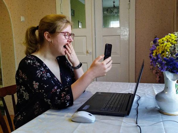 Yawning european young woman sit at table and have video call on smartphone at home - Fotó, kép