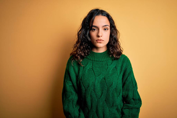 Young beautiful woman with curly hair wearing green casual sweater over yellow background with serious expression on face. Simple and natural looking at the camera. - Fotografie, Obrázek