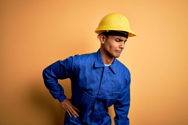 Jeune beau travailleur afro-américain portant un uniforme bleu et un casque de sécurité Souffrant de maux de dos, touchant le dos avec la main, douleurs musculaires
 - Photo, image