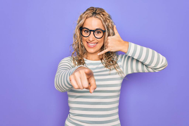 Beautiful blonde woman wearing casual striped t-shirt and glasses over purple background smiling doing talking on the telephone gesture and pointing to you. Call me. - Photo, Image