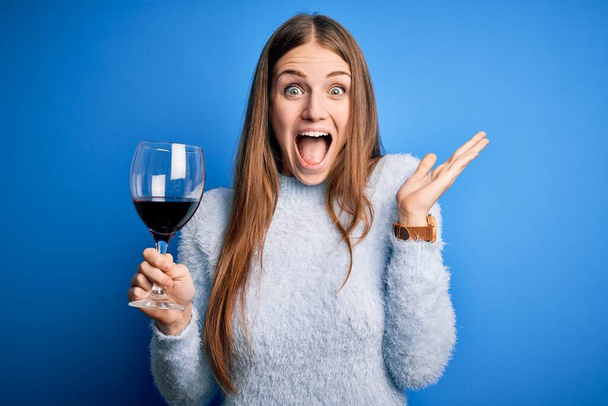 Young beautiful redhead woman drinking glass of red wine over isolated blue background very happy and excited, winner expression celebrating victory screaming with big smile and raised hands - Photo, Image