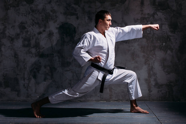 young strong guy, karate in a white kimono in the studio on a black background performs a punch - Foto, imagen