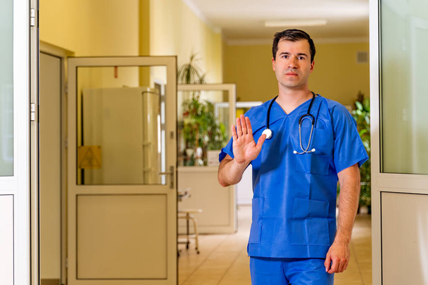 Middle age, Caucasian white, male doctor in scrubs in hospital corridor, showing stop with palm. Stop sign for coronavirus or covid 19 - Photo, Image