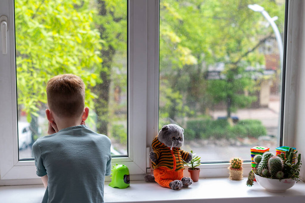 Мальчик сидит на подоконнике и смотрит на прекрасную игрушечную кошку. Концепция Stay at home во время пандемии covid-19 - Фото, изображение