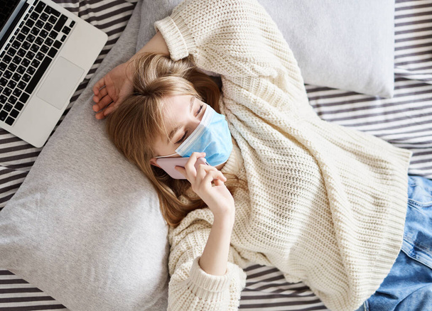 Beautiful teenager in medical mask wearing white sweater and jeans talking via cell phone lying on a bed with laptop nearby. Young girl wearing protective mask and making a call. - Φωτογραφία, εικόνα