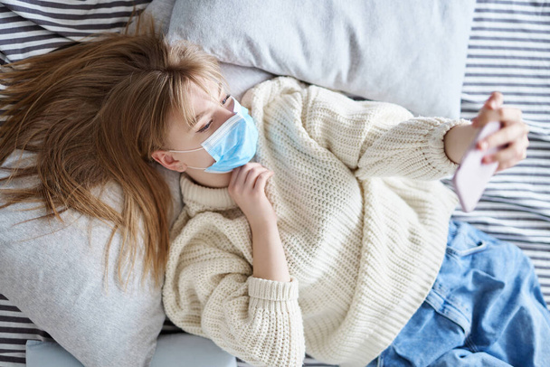 Selfie concept. Smiling beautiful young woman in protective medical mask in sweater and jeans lying on a bed and makes self-portrait by smartphone. Quarantine or isolation during coronavirus covid-19. - Photo, Image