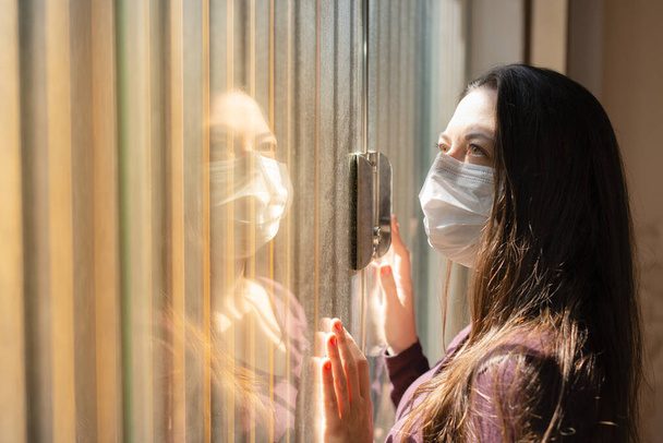 A woman under quarantine at home, wearing a medical face mask and a casual outfit, is standing idle in front of a closed window, the hand on the glass, staring into space. Stay at Home Concept. - Photo, Image