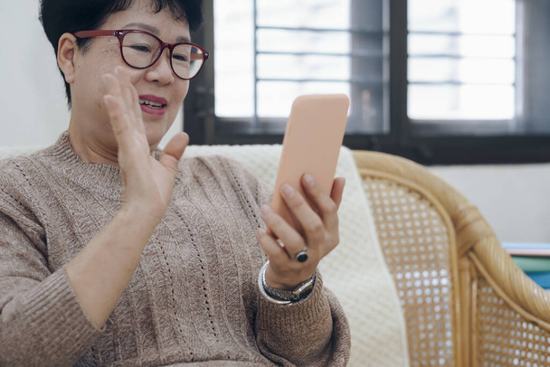 Aasialainen vanhempi nainen, joka soittaa videopuhelun matkapuhelimella, vilkuttaa näytöllä, chattailee verkossa etäisten ihmisten kanssa
. - Valokuva, kuva