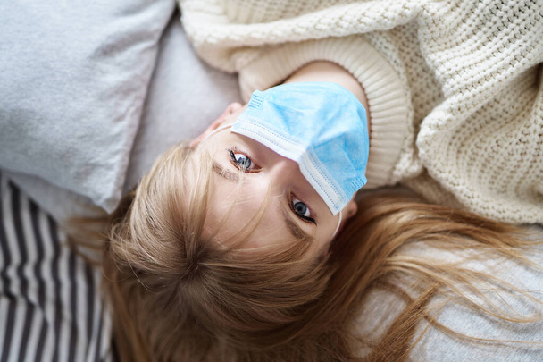Ein junges attraktives blondes Mädchen in weißem Pullover, das mit einer medizinischen Maske auf einem Bett liegt und direkt in die Kamera blickt. Coronavirus, Quarantäne oder Isolationskonzept. - Foto, Bild