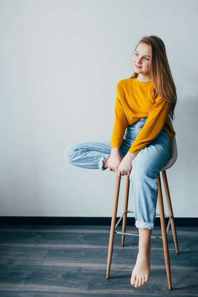 teenage girl looks at left side sitting on white modern chair with one  leg raise up. Beautiful girl in yellow sweater and blue jeans in home interior with black wooden floor. Trendy casual outfit.  - Photo, image