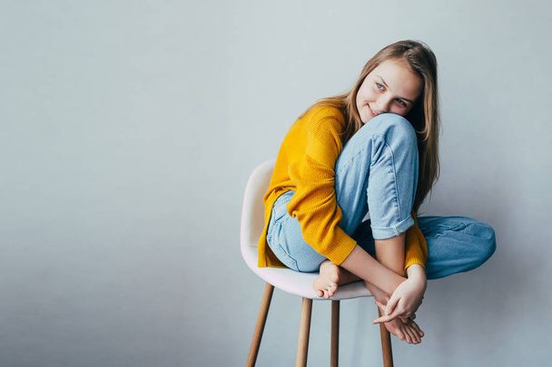 teenage girl looks at side and sitting on white modern chair with legs up. Beautiful girl in yellow sweater and blue jeans in home interior. Trendy casual outfit. place for text - Fotó, kép