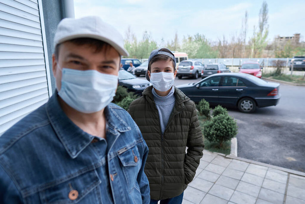 мужчина и подросток позируют возле стены и закрытой двери высотных зданий с квартирами, жилым районом, медицинской маской на лицах защищает от вирусов и пыли
 - Фото, изображение