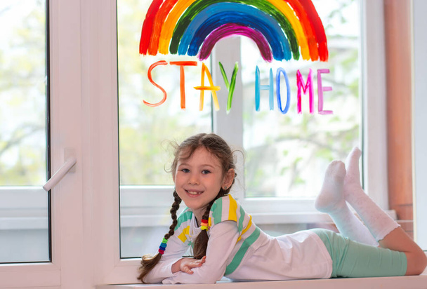Šťastná dívka leží na parapetu na pozadí okna s nápisem "zůstaň doma" a duhou malovanou barvami. Karanténní zábava. - Fotografie, Obrázek