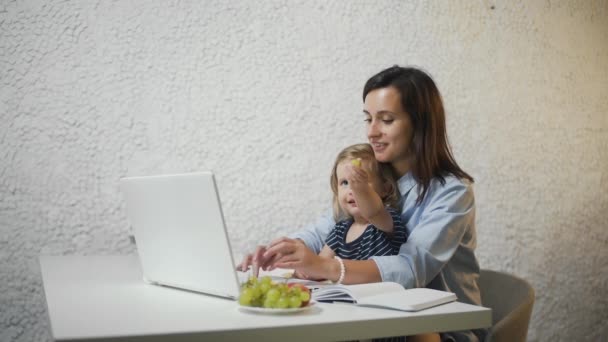 Молодая мать работает на ноутбуке, пока ее ребенок сидит на коленях и ест виноград
 - Кадры, видео