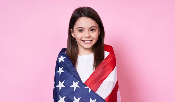 Cute stylish young emotional girl, long-haired brunette, smiles, covered herself with the American flag, Independence Day, July 4, victory,peace,USA, in a white base T-shirt on a pink background. - Photo, Image