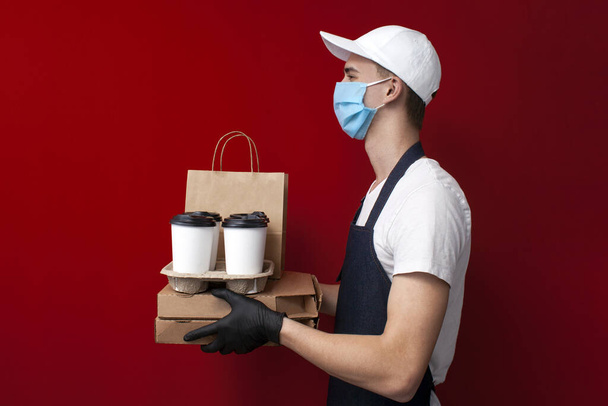 young professional food delivery man in a protective mask and gloves holds boxes of pizza and coffee and gives the client, fast delivery during the epidemic of coronavirus - Photo, Image