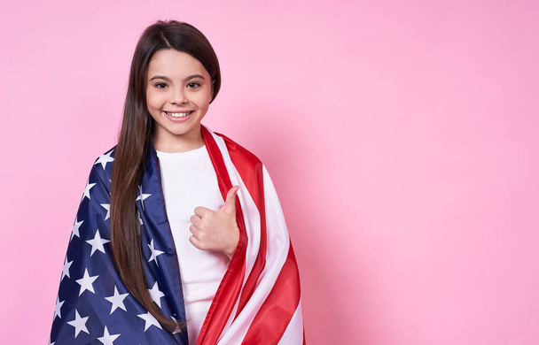 Happy stylish young emotional girl, long-haired brunette, smiles broadly and holds joyfully American flag, Independence Day, July 4,peace,victory, USA,like,ok.In abase T-shirt on a pink background. - Zdjęcie, obraz