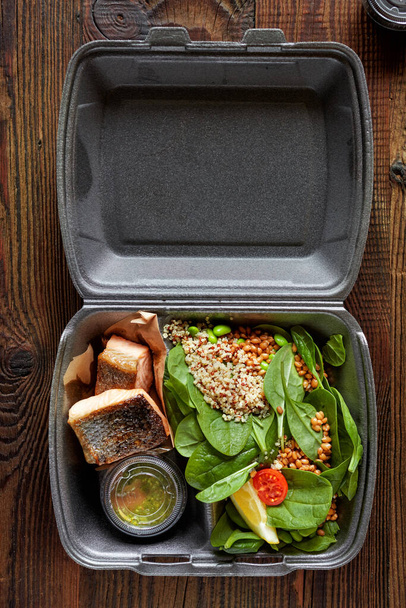 fried salmon pieces and spinach salad in plastic take away box on wooden table, food delivery - Foto, afbeelding