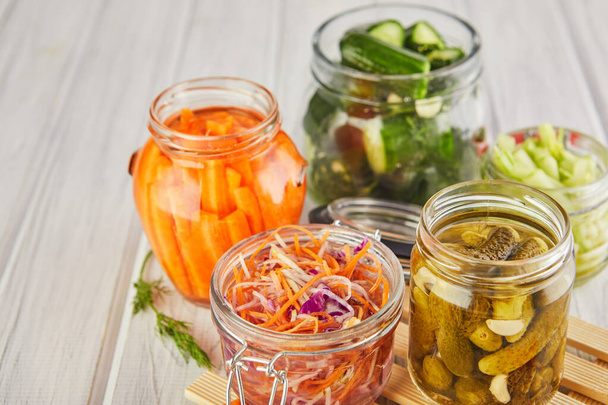 Fermented preserved vegetarian food concept. Sour sauerkraut, pickled carrots, pickled cucumbers, pickled celery in glass jars on a white wooden kitchen table. The concept of canned food. Copy space - Photo, Image