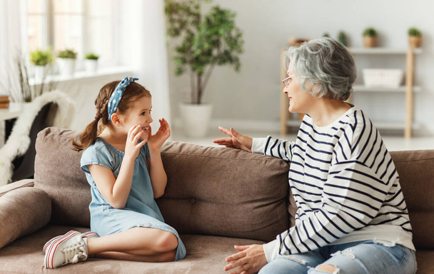 Little girl smiling and talking with astonished elderly woman while sitting on sofa at home togethe - Photo, Image