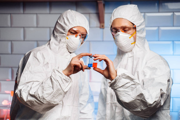 コロナウイルスワクチン開発。防護服、マスク及びガラスの研究者は、フラスコ及び試験管を用いて化合物を合成し、液体を探す実験を行う。. - 写真・画像
