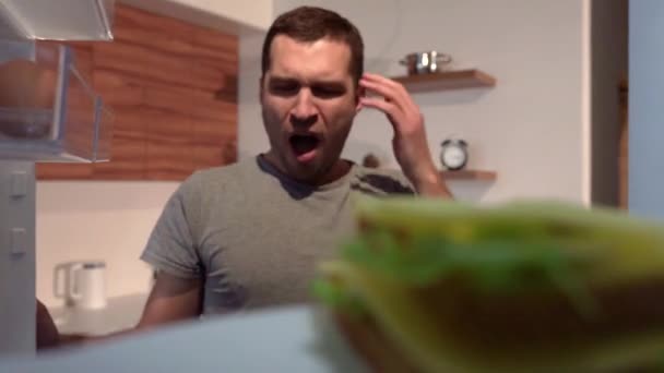 Video of young sleepy man opened fridge door and take bitten sandwich to take a bite. Chewing delicious snack and put it back on plate and close fridges door. - Imágenes, Vídeo