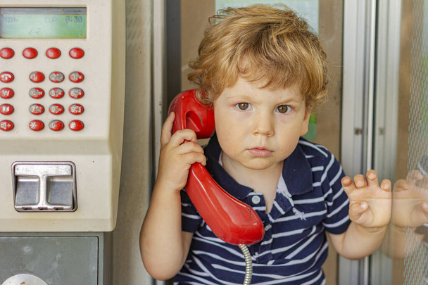 Érzelmes gyerek beszél a telefonban. A fiú a telefonfülkében. Egy kisfiú piros telefonnal a kezében. - Fotó, kép