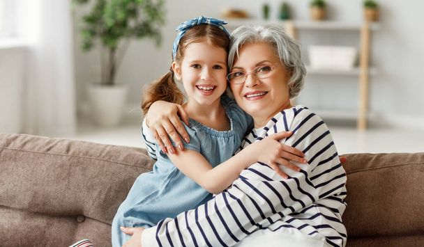 Joyful little girl hugging happy grandmother   while sitting together on sofa and looking at camera on grandparents da - Фото, изображение