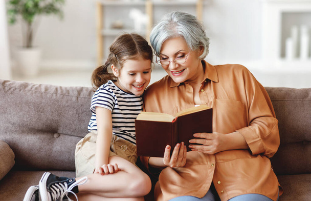 Счастливая пожилая женщина и маленькая девочка улыбаются, сидя на диване и читая увлекательную сказку вместе в доме
 - Фото, изображение