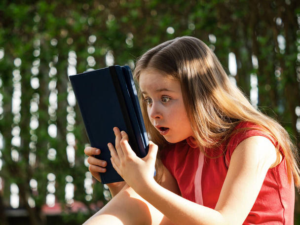 Distance learning during the quarantine period for the virus Covid-19. Cute little schoolgirl with long hair is studying from home, sitting in the garden on the grass. Uses a tablet and remote work via the Internet. - Fotoğraf, Görsel