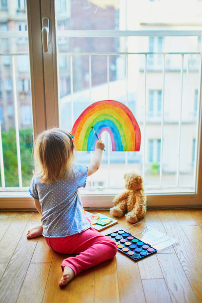 Roztomilé batole dívka malování duha na okenní sklo jako znamení naděje. Tvůrčí hry pro děti, které zůstávají doma během uzamčení. Samostatná izolace a koncepce karantény koronaviru - Fotografie, Obrázek