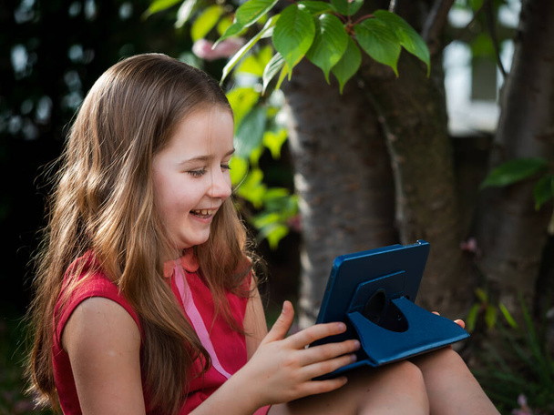 Distance learning during the quarantine period for the virus Covid-19. Cute little schoolgirl with long hair is studying from home, sitting in the garden on the grass. Uses a tablet and remote work via the Internet. - Zdjęcie, obraz