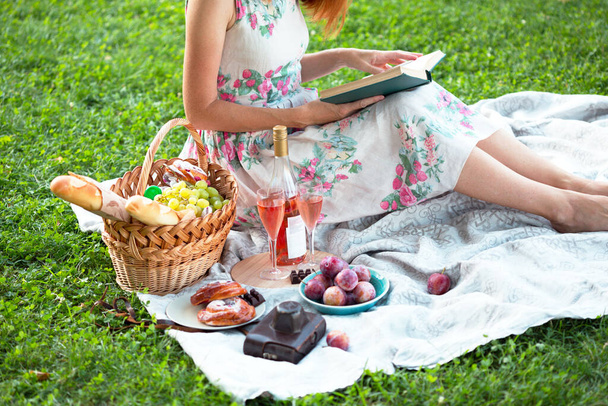 Summer - picnic in the meadow.  girl sitting reading a book and near a picnic basket and baguette, wine, glasses, grapes and roll - Fotó, kép