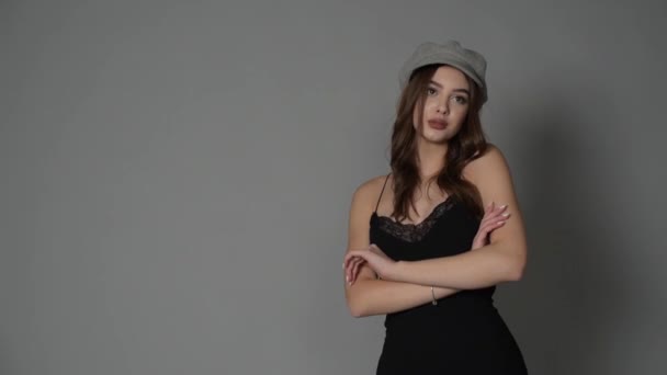 Glamorous young woman wears fashionable dress posing with fluttering hair at grey wall. Girl put forward hand in a black dress and a gray cap. Beautiful young girl with brown curly hairs. portrait - Footage, Video
