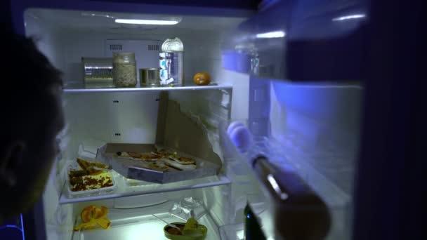 Een man op zoek naar eten in de koelkast. Een ongeschoren man met een fles bier in zijn hand is op zoek naar iets eetbaars in de koelkast. Nachthonger. Een kater. Vrijgezellenkoelkast, vol muf eten. - Video