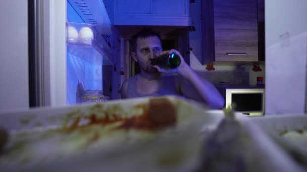 Man looking for food in the fridge. An unshaven man with a bottle of beer in his hand is looking for something edible in the fridge. Night hunger. Hangover. Bachelor's fridge, full of stale food. - Footage, Video