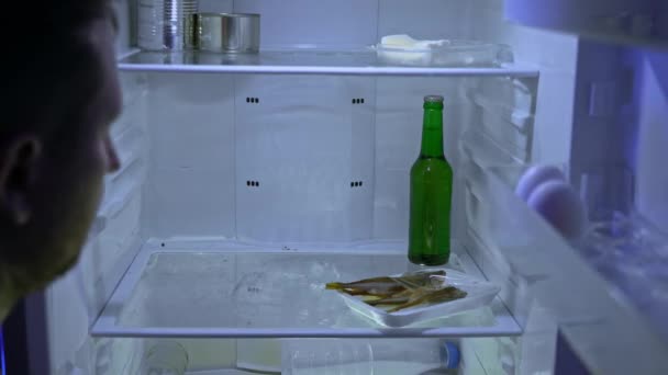 Man looking for food in the fridge. The guy sniffs fish from the fridge, takes it and a bottle of beer. Night hunger. Hangover. Bachelor's fridge, view from behind. Dirty fridge. - Footage, Video