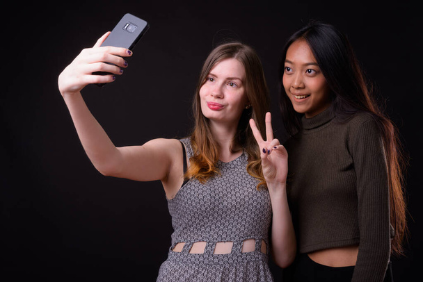Studioaufnahme der jungen schönen Asiatin und der jungen schönen Skandinavierin zusammen vor schwarzem Hintergrund - Foto, Bild