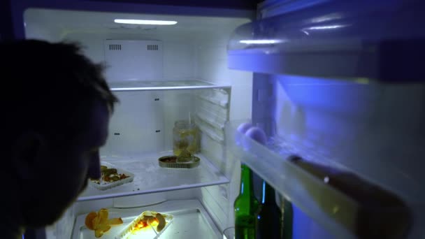 Ein Mann in der Isolation sucht nach Nahrung im Kühlschrank. Essensreste im Kühlschrank während der Quarantäne. Der unrasierte Mann wühlt in seinem Kühlschrank. Ungesunde Ernährung. Nächtlicher Hunger. Katerstimmung. Bachelor 's Kühlschrank. Schmutziger Kühlschrank voller abgestandener Lebensmittel. - Filmmaterial, Video