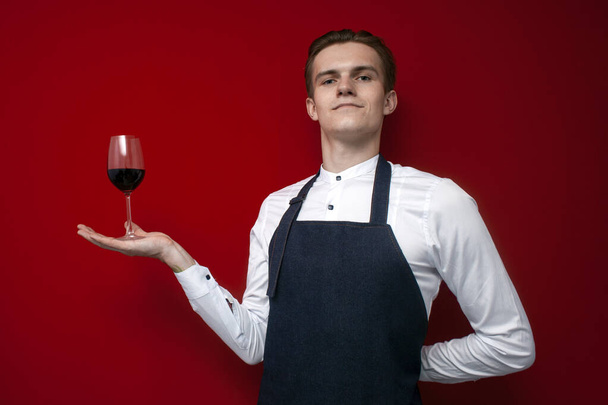 joven camarero profesional en uniforme sostiene una copa de vino tinto y sonriendo sobre un fondo rojo, un tipo sommelier recomienda vino
 - Foto, Imagen