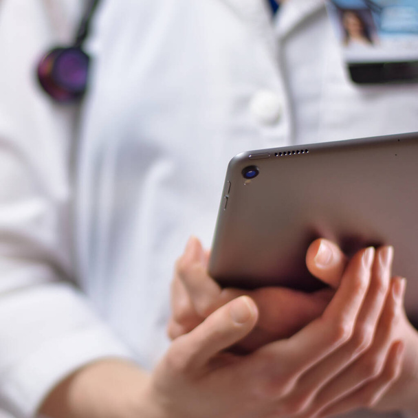 Tablet in the hands of healthcare professional up close. White coat, stethoscope, and badge visible in background. Hands of nurse practitioner or PA using technology in medicine for patients - Photo, Image