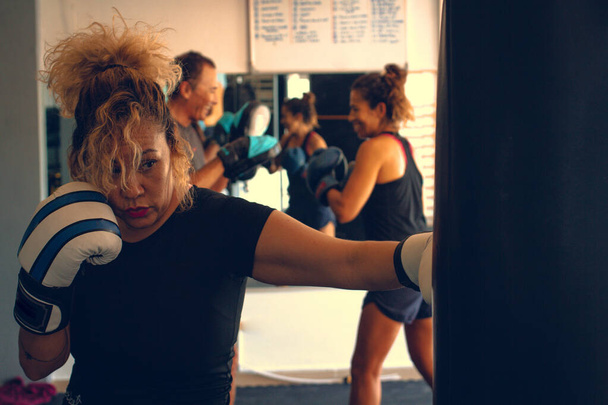 Blondýny žena cvičit boxerské bodnutí s modrými a bílými rukavicemi punching boxerský pytel v tělocvičně - Fotografie, Obrázek